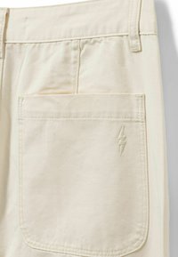 Cream-colored pants featuring a single back pocket with a stitched lightning bolt detail, made from a soft cotton blend fabric.