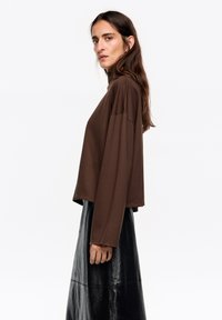 Brązowa, luźna bluzka z długim rękawem i wysokim dekoltem, zestawiona z błyszczącą czarną skórzaną spódnicą. Gładka tekstura, casualowy styl.