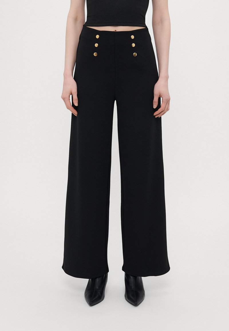 Pantalon noir taille haute à jambes larges avec détails de boutons dorés, porté avec des bottines noires à bout pointu et un haut court noir.