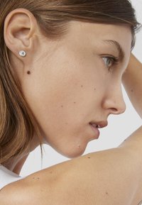 Orecchino a perno in acciaio inossidabile chirurgico con una pietra rotonda di zirconia cubica trasparente, indossato all'orecchio sinistro di una persona con tono di pelle medio.