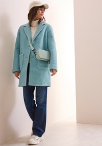Cappotto azzurro chiaro, tessuto strutturato con grandi tasche; dolcevita crema; jeans blu; sneakers bianche; piccola borsa a tracolla azzurro chiaro; cappello crema.
