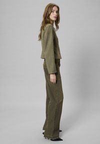 Kruvi rohelise värvi lühike jakk tekstureeritud kangast, millele on kombineeritud kõrge vöökohaga laiad oliivirohelised püksid, mille allääres on frantsi ning peenikese musta jalanõudega.