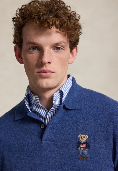 Νεαρός άνδρας με μπούκλες κόκκινα μαλλιά, φοράει μπλε πλεκτή μπλούζα πόλο με μικρή κεντημένη αρκουδάκι που κρατάει σημαία στο στήθος.