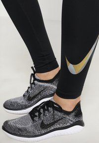 Černé sportovní legíny v kombinaci s černobílými běžeckými teniskami Nike, které mají texturovaný svršek a šedé a zlaté akcenty.