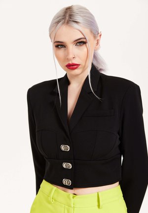 Black cropped blazer made of smooth fabric, featuring a tailored design, three front buttons, and two chest pockets. Neon green pants visible.