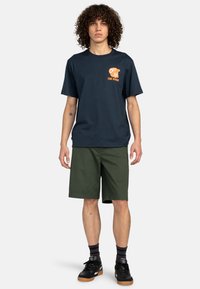 Dunkelblaues Baumwoll-T-shirt mit orangefarbener Grafik auf der Brust, kombiniert mit grünen Shorts. Schwarze Sneakers mit Gummisohle vervollständigen das Outfit.
