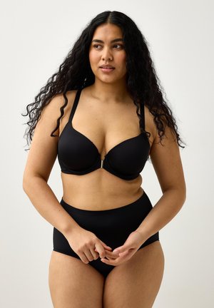 Kvinde med langt krøllet hår iført en sort bh og sort højtaljet undertøj, der kigger til siden med hænderne løst foldet.