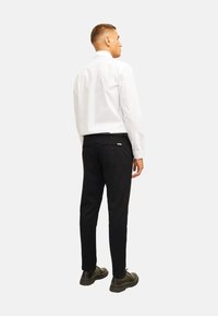Camisa blanca de manga larga con collar clásico, combinada con pantalones negros ajustados y zapatos oscuros. Presenta un diseño minimalista y detalles sutiles.