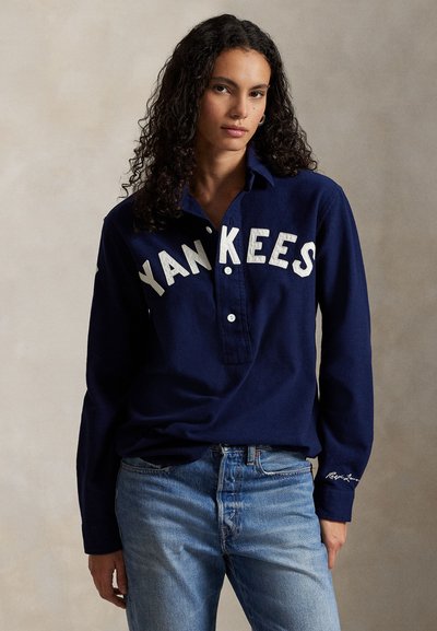 Camisa polo de mangas longas na cor azul-marinho com "YANKEES" bordado em branco ao longo do peito, apresentando um colarinho com botões e um corte descontraído.