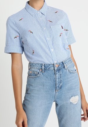 Kvinde iført en kortærmet blå og hvid stribet skjorte med knapper og broderede fugle, stoppet ned i højttaljede lyseblå revnede jeans.