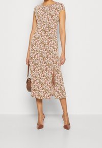 Robe midi à motif floral rose, blanc et vert, manches courtes et fente latérale. Portée avec des talons marron et un sac assorti.