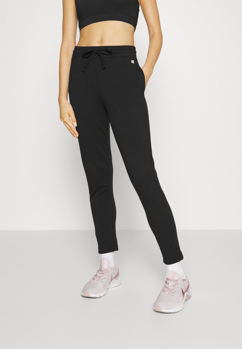 Jogging en coton noir avec taille ajustable par cordon de serrage, ourlets élastiqués et poches latérales. Porté avec des chaussures de sport rose clair.