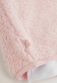 Gaiši rozā flīsa sega ar mīkstu, teksturētu virsmu; stūrī ir iešūts neliels zīmola etiķete.