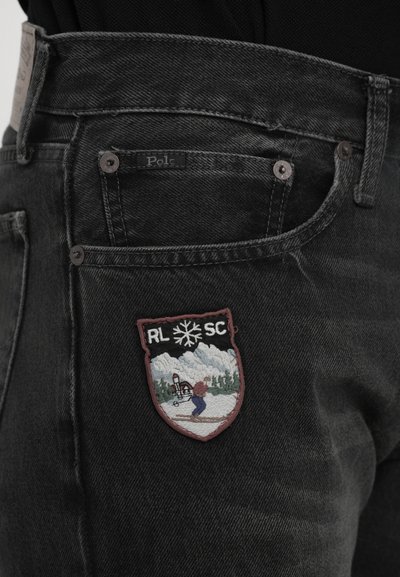 Mustharjadest teksapüksid, mille taskul on tikandiga patch, kujutades suusatajat, lumiseid mägesid, puid ja teksti "RL SC" koos lumehelvesümboliga.