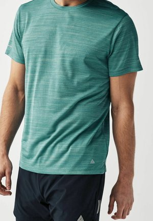 Homme portant un t-shirt de sport bleu sarcelle à manches courtes et un short noir, visible du cou jusqu'au milieu de la cuisse sur un fond uni.