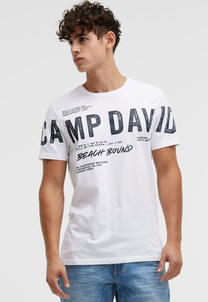 Junger Mann mit lockigem Haar, trägt ein weißes "CAMP DAVID"-T-Shirt mit Grafik und blaue Jeans, schaut nach links vor einem einfarbigen Hintergrund.