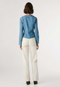 Sieviete stāv ar muguru vērsta, ģērbta zilu džinsu jaku, krēma platām biksēm un melniem augstpapēžu apaviem uz vienkārša fona.