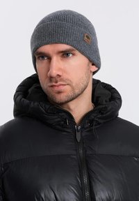 Beanie grigia a coste con una toppa in pelle marrone, indossata con un giubbotto imbottito nero dotato di colletto alto e chiusura con zip.