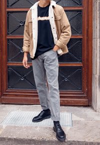 Beżowa kurtka z białym futrzanym kołnierzem, czarny t-shirt z grafiką pod spodem, szare spodnie i czarne sznurowane buty, stojąc na teksturowanej powierzchni.