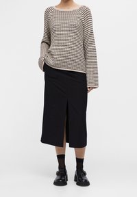 Persoon in een beige en zwart gestreepte lange mouw top, zwarte midi rok met een split aan de voorkant, zwarte sokken en zwarte schoenen met gesp, staand op een witte vloer.