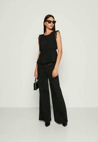 Black lace-accented top with peplum waist, paired with high-waisted black wide-leg trousers. Model holds a small black handbag.