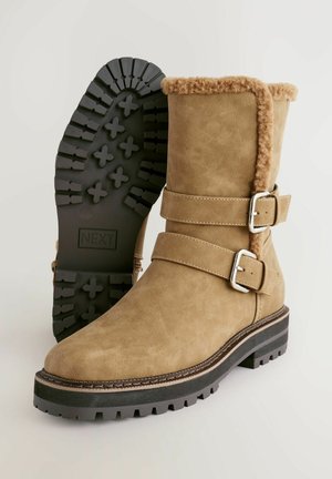 Stivaletti alla caviglia in suede marrone con bordo in pelliccia sintetica, due fibbie argentate e suola in gomma nera texturizzata con battistrada decorato.