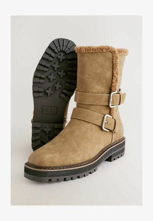 Brūni zamša potītes zābaki ar mākslīgās kažokādas apdari, diviem sudraba sprādzēm un strukturētu melnu gumijas zoli ar rakstainu profilu.