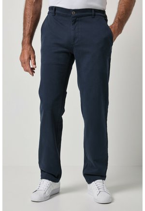 Hombre con pantalones azul marino de corte recto con bolsillos delanteros y zapatillas blancas, de pie frente a un fondo liso.