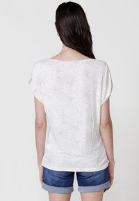 Haut à manches courtes blanc avec un motif floral dans une teinte beige clair ; coupe décontractée, encolure arrondie et style d'ourlet casual, associé à un short en jean.