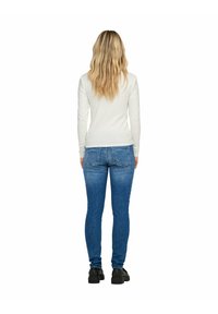 Weißes Langarmoberteil, kombiniert mit blauen Skinny-Jeans, die eine glatte Baumwolltextur und eine klassische Passform aufweisen. Schwarze Schuhe vervollständigen das Outfit.