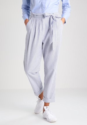 Kobieta w jasnoniebieskich, przewiewnych spodniach z wysokim stanem i paskiem do wiązania, w połączeniu z jasnoniebieską koszulą i białymi trampkami.