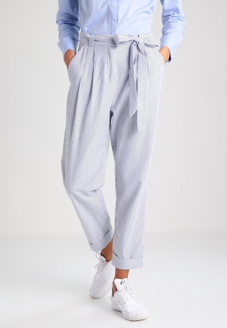 Femme portant un pantalon taille haute bleu clair à fines rayures avec une ceinture nouée, associé à une chemise bleu clair et des baskets blanches.