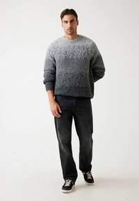 Sivý sveter s prechodovým zdobením a textúrovaným povrchom, kombinovaný s tmavými džínami a čiernymi teniskami s bielymi šnúrkami. Model stojí sebavedome.