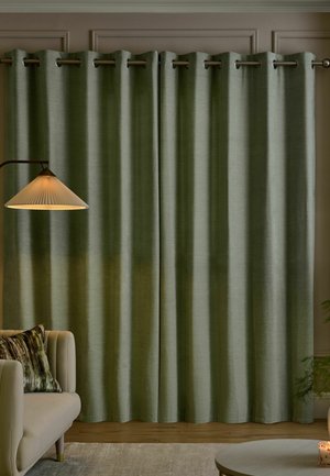 Grüne Vorhänge, die an einer Metallstange hinter einem beigen Sofa mit Kissen hängen, neben einer beleuchteten Stehlampe und einem runden Tisch mit einer Kerze.