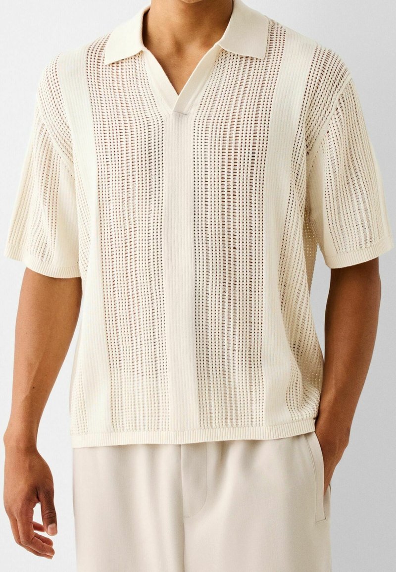 Polo de punto en crema claro con mangas cortas, que presenta un patrón de calado y acentos de canalé en el cuello y el dobladillo.