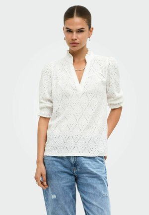 Femme portant un chemisier en dentelle blanche à motifs ajourés avec un col à volants et des manches retroussées, associé à un jean bleu clair déchiré.