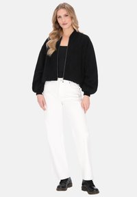 Chaqueta bomber negra con diseño texturizado, cierre de cremallera y mangas anchas, combinada con pantalones anchos blancos y botas negras.