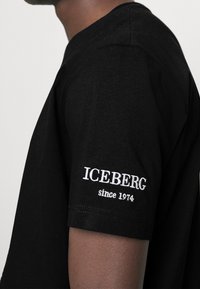Black cotton t-shirt featuring a crew neck and short sleeves. White embroidered "ICEBERG since 1974" on the left sleeve. Smooth texture.