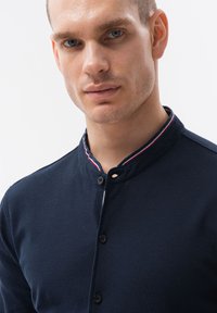 Tummansininen napillinen paita kauluksella, jossa on punavalkoisia raidallisia yksityiskohtia ja teksturoitu kangas. Säädettävä nappikiinnitys.