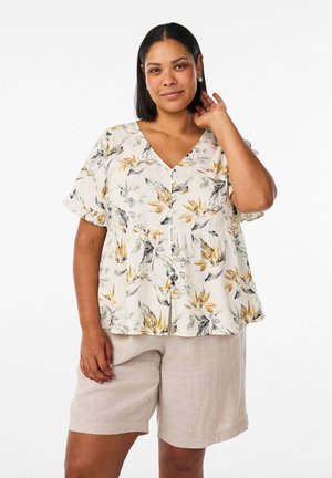 Kvinde med mellemtonet hud og glat, sort hår iført kortærmet blomstret bluse og beige knælange shorts, stående foran en hvid baggrund.