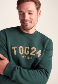 Green sweatshirt made of soft fabric, featuring a round neckline and large gold textured print "TOG24 EST. 1958" on the front.