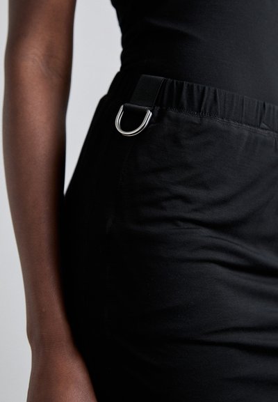 Îmbrăcăminte din material elastic negru cu un tiv elastic, având o bandă neagră cu un detaliu din inel D metalic pe lateral. Textură netedă.