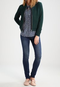Grüne gesteppte Jacke mit silbernem Reißverschluss, blumige Bluse, blaue Skinny Jeans und hellrosa Slipper mit dekorativen Akzenten.