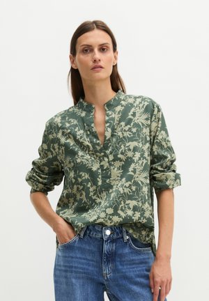 Vrouw draagt een groene bloemen lange mouwen blouse met opgerolde mouwen en blauwe jeans, staand met één hand in de broekzak.