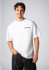 Weißes Kurzarm-T-Shirt mit dem Aufdruck "DOWNTOWN CLUB" in Schwarz auf der linken Brust. Glattes Baumwollmaterial, lässige Passform, Rundhalsausschnitt.