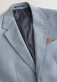 Blazer bleu clair texturé avec doublure intérieure noire et un mouchoir de poche marron dans la poche poitrine, posé à plat sur une surface blanche.