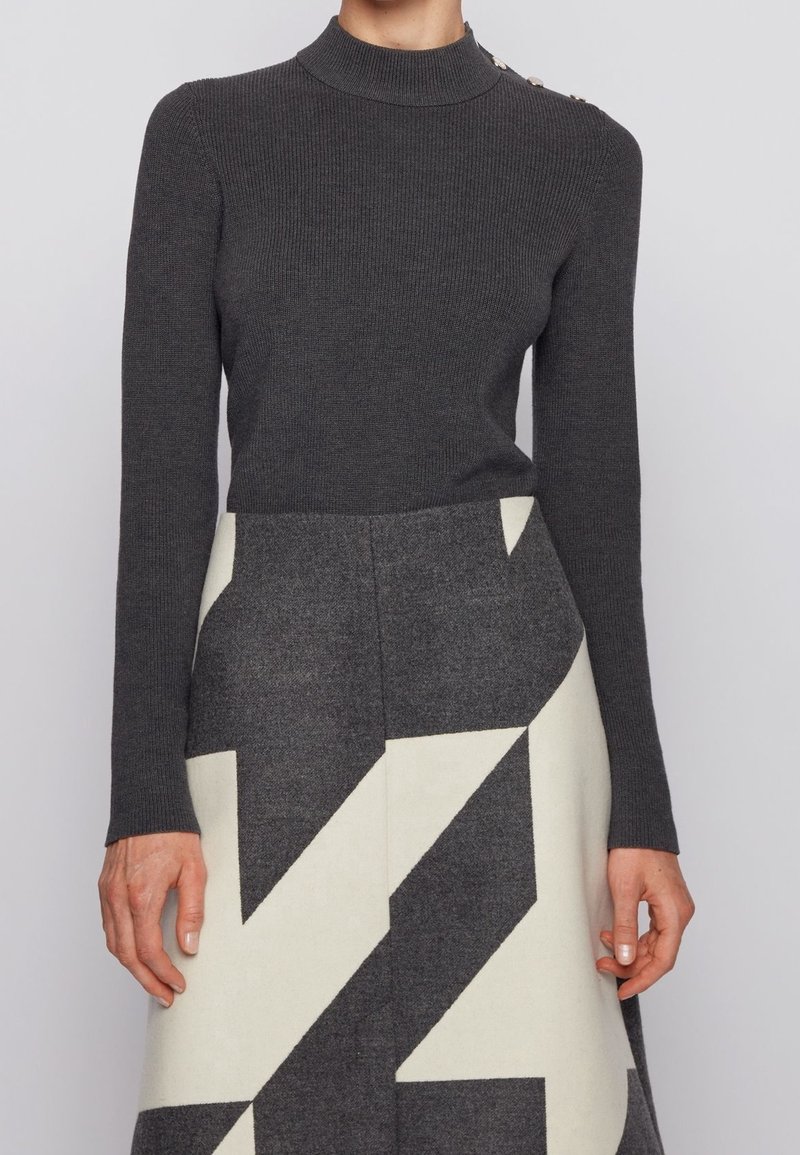 Pull à col roulé côtelé gris foncé à manches longues, associé à une jupe longueur genou ornée de motifs géométriques audacieux en gris et blanc cassé.