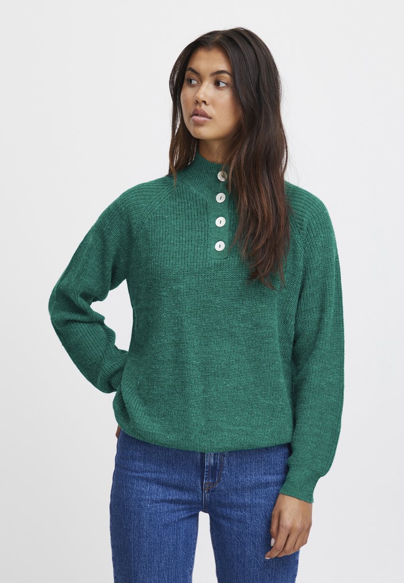 Groene gebreide trui met een hoge hals, vier witte knopen en geribbelde textuur. Gekocht met blauwe denim jeans.