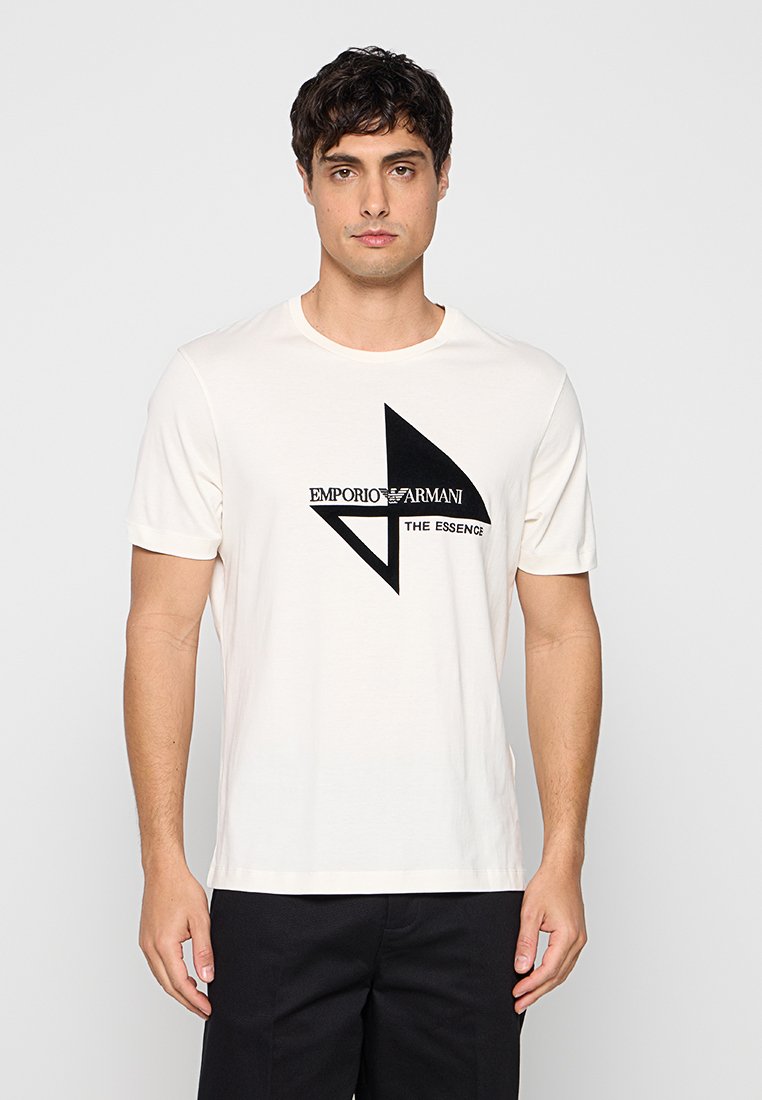 Hombre que lleva una camiseta blanca de manga corta con diseño geométrico negro y texto "EMPORIO ARMANI THE ESSENCE", combinada con pantalones negros.