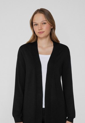 Cardigan negro abierto por delante con mangas largas y una tela lisa y texturizada. Se lleva sobre una blusa blanca con cuello redondo.